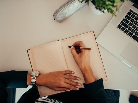 Hände notieren etwas in ein offenes Notizbuch neben einem aufgeklappten Laptop