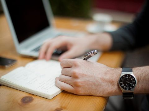Hand mit Armbanduhr notiert Punkte in ein Notizbuch neben einem Laptop