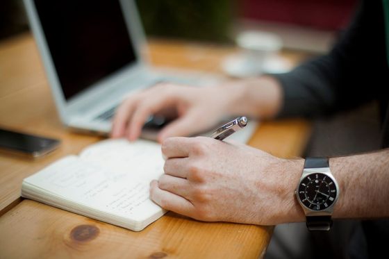 Hand mit Armbanduhr notiert Punkte in ein Notizbuch neben einem Laptop